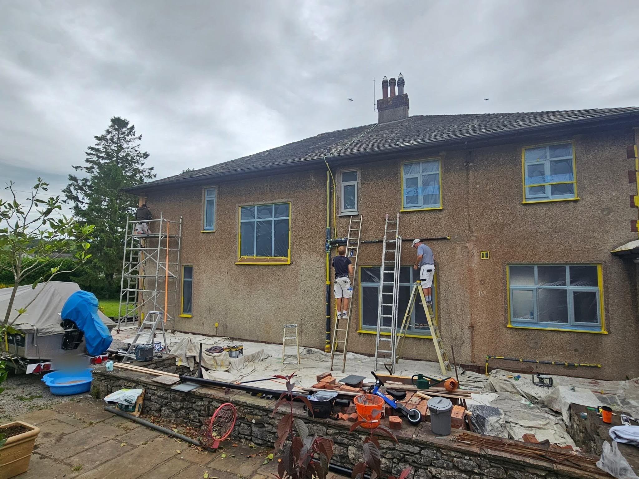 Two decorators on ladders preparing a house, windows masked in yellow tape and dust sheets down